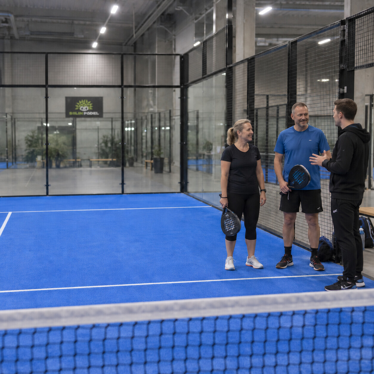 Ohjaaja neuvoo kahta aloittelijaa padelkentällä alkeiskurssilla